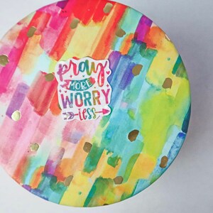 May include: A round, colorful container with a watercolor design featuring streaks of pink, orange, yellow, green, and blue. Gold glitter dots are scattered across the surface. A white sticker with the text "pray more worry less" is centered on the container.
