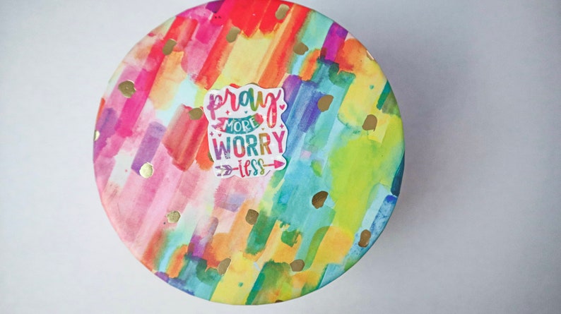 May include: A round, colorful container with a watercolor design featuring streaks of pink, orange, yellow, green, and blue. Gold glitter dots are scattered across the surface. A white sticker with the text "pray more worry less" is centered on the container.