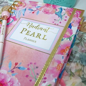 May include: A pink and white floral planner with gold accents and the text "Radiant PEARL PLANNER". A white pen with a pink top is lying on top of the planner.
