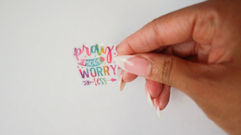 May include: A colorful sticker with the text "pray more worry less" in a rainbow font. The sticker has a white background and a rainbow arrow pointing to the right.
