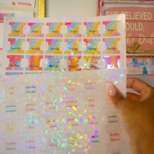 May include: A sheet of iridescent rainbow holographic stickers with words like "peace", "love", "gratitude", and "joy" printed on them. The stickers are arranged in rows and columns on a white background.
