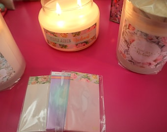 May include: Three glass candle jars with floral designs and the text "You Are Radiant" on the labels. The candles are on a pink surface with a set of floral patterned notepads.
