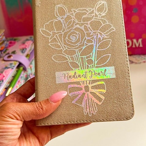May include: A beige notebook with a floral design and a holographic sticker that says "Radiant Pearl".