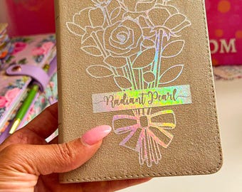 May include: A beige notebook with a floral design and a holographic sticker that says "Radiant Pearl".