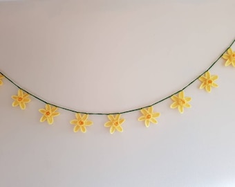May include: A garland of crocheted yellow flowers with orange centers strung on a green thread. The flowers are evenly spaced and appear to be daffodils. The garland is hanging against a plain white wall.
