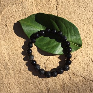 May include: Black beaded bracelet with black lava rock beads. The bracelet is on a green leaf.