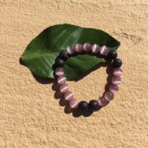 May include: A bracelet made with pink and black beads. The pink beads are round and have a shimmery finish. The black beads are round and have a rough texture. The bracelet is lying on a green leaf.
