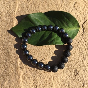 May include: A black beaded bracelet with alternating smooth and textured beads. The bracelet is resting on a green leaf.