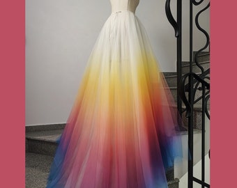 May include: A full-length, multi-layered tulle skirt on a mannequin. The skirt transitions from white at the waist to a vibrant gradient of yellow, orange, red, purple, and blue at the hem. The skirt is displayed on a mannequin in front of a staircase.