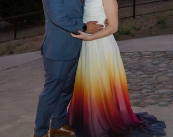 May include: A couple poses for a wedding photo. The groom is wearing a blue suit and a tie. The bride is wearing a white dress with a gradient of yellow, orange, red, and purple. The bride has blue hair and is wearing a flower in her hair.