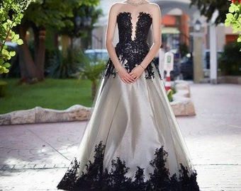 May include: A woman in a strapless, silver satin gown with black lace detailing. The gown has a fitted bodice and a full skirt. She is standing in front of a green archway with red flowers.