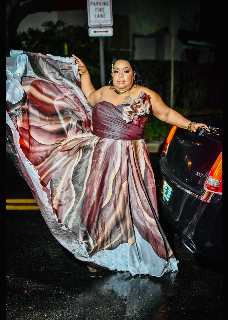 Peut inclure: Robe bustier aux tons bordeaux, marron et cr&egrave;me, avec une jupe fluide. La robe est orn&eacute;e d'un motif floral et est port&eacute;e &agrave; l'ext&eacute;rieur pr&egrave;s d'une voiture noire. Le tissu semble l&eacute;ger et a&eacute;r&eacute;.
