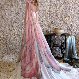 Peut inclure: Une longue robe fluide avec un motif aquarelle rose, blanc et bleu. La robe pr&eacute;sente une superposition transparente avec un motif floral et une longue tra&icirc;ne. Le dos de la robe est visible, avec des d&eacute;tails de bretelles d&eacute;licats.