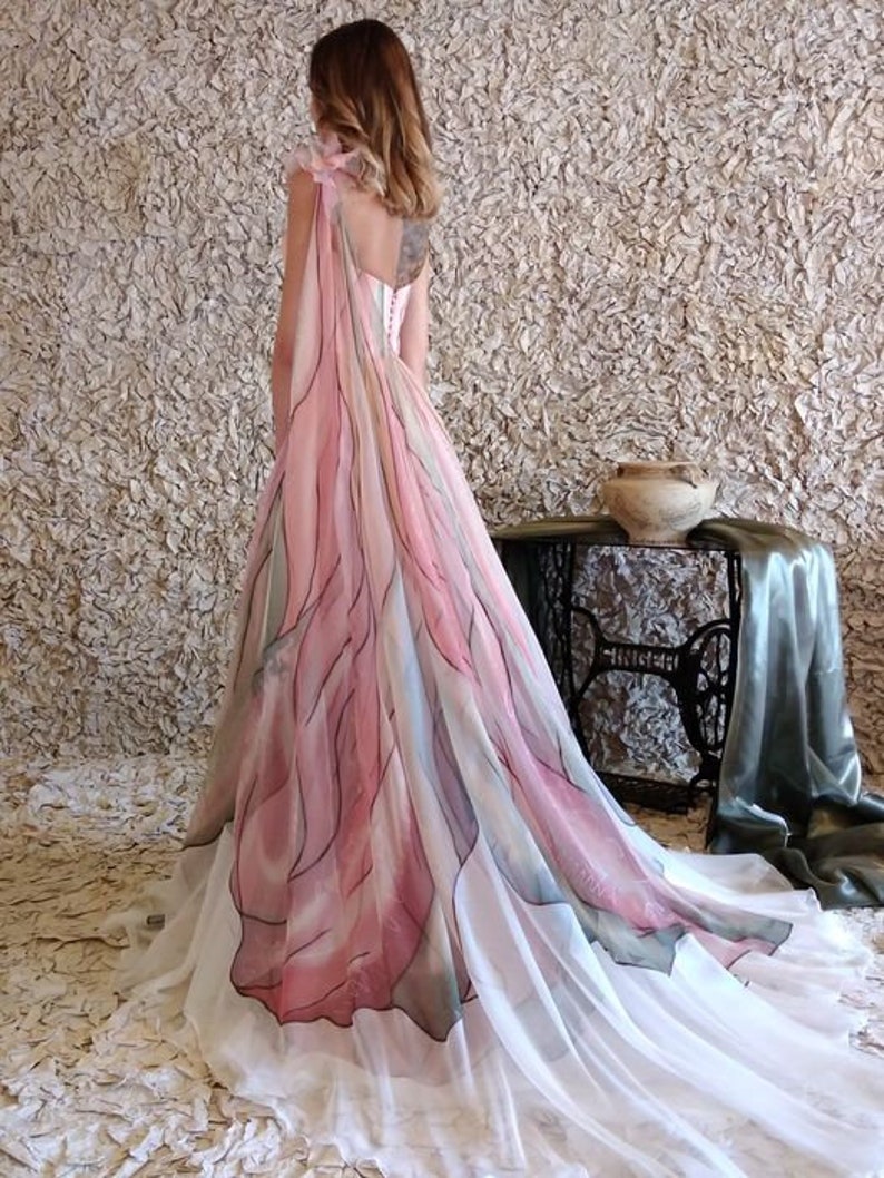 Peut inclure: Une longue robe fluide avec un motif aquarelle rose, blanc et bleu. La robe pr&eacute;sente une superposition transparente avec un motif floral et une longue tra&icirc;ne. Le dos de la robe est visible, avec des d&eacute;tails de bretelles d&eacute;licats.