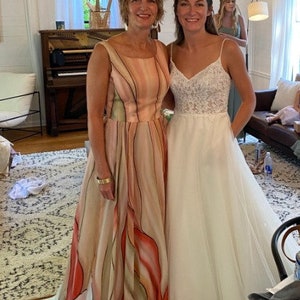 Peut inclure: Deux femmes posent &agrave; l'int&eacute;rieur. L'une porte une longue robe fluide avec un motif floral dans les tons p&ecirc;che, rouge et blanc. L'autre porte une robe de mari&eacute;e blanche avec un corsage en dentelle et des bretelles spaghetti. Un lustre est suspendu au-dessus.