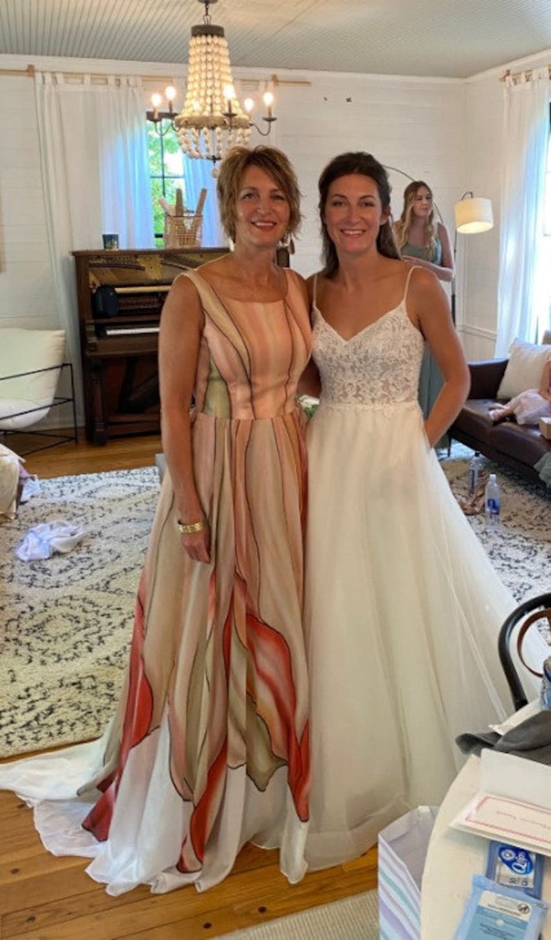 Peut inclure: Deux femmes posent &agrave; l'int&eacute;rieur. L'une porte une longue robe fluide avec un motif floral dans les tons p&ecirc;che, rouge et blanc. L'autre porte une robe de mari&eacute;e blanche avec un corsage en dentelle et des bretelles spaghetti. Un lustre est suspendu au-dessus.