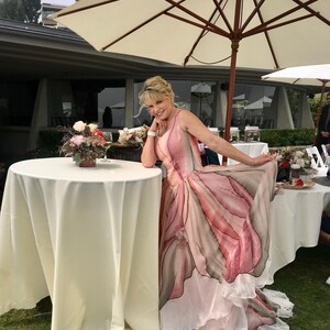 Peut inclure: Une femme porte une robe &agrave; imprim&eacute; floral rose et gris, posant pr&egrave;s d'une table recouverte d'une nappe blanche. La robe pr&eacute;sente une jupe &agrave; plusieurs &eacute;paisseurs et un motif unique en forme de p&eacute;tale. Un grand parasol cr&egrave;me offre de l'ombre.
