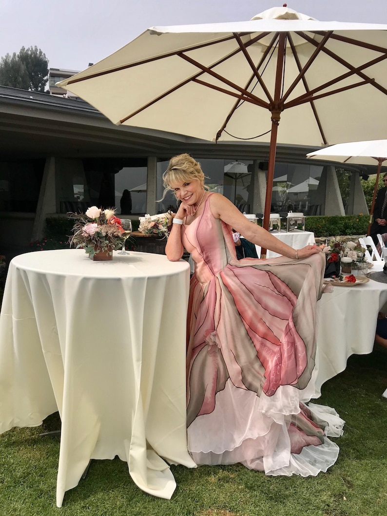 Peut inclure: Une femme porte une robe &agrave; imprim&eacute; floral rose et gris, posant pr&egrave;s d'une table recouverte d'une nappe blanche. La robe pr&eacute;sente une jupe &agrave; plusieurs &eacute;paisseurs et un motif unique en forme de p&eacute;tale. Un grand parasol cr&egrave;me offre de l'ombre.