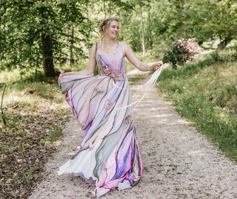 Peut inclure: Robe de mari&eacute;e sans manches et fluide avec un motif aquarelle dans les tons violets, roses et verts. La robe pr&eacute;sente des ornements floraux &agrave; la taille et une longue jupe fluide. La mari&eacute;e tient un bouquet de fleurs roses.