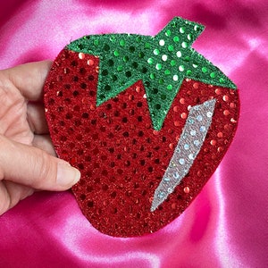 Puede incluir: Un parche de fresa rojo y verde con lentejuelas y un diseño de purpurina plateada.