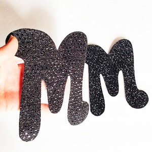 May include: Two black glitter letters, both shaped like the letter "M", are shown. The letters are slightly overlapping.