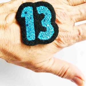 May include: A black felt patch with the number 13 in blue glitter. The patch is on a person's hand.