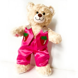Puede incluir: Un oso de peluche de peluche que lleva un atuendo de satén rosa brillante con aplicaciones de fresas. El atuendo incluye un chaleco y pantalones, con detalles plateados. El oso tiene pelaje marrón claro y una expresión amigable.