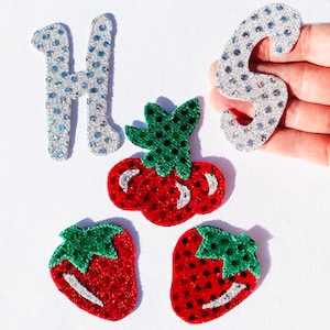 Puede incluir: Un conjunto de cuatro parches para planchar con un diseño de fresa roja y verde y dos letras de purpurina plateada, "H" y "S".