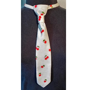May include: A light gray tie with a repeating pattern of red cherries and green leaves. The tie is knotted and displayed against a dark background. The tie has a ribbed texture.