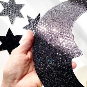 May include: A black and silver glitter crescent moon and three black glitter stars. The moon has a gradient of glitter, with the silver glitter at the top and the black glitter at the bottom.