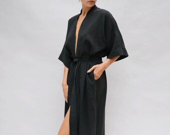 May include: A black linen robe with a wrap-around design and a matching belt. The robe features wide sleeves and a knee-high slit on one side. The model is wearing the robe, standing against a plain white background.