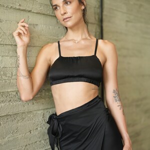 May include: A black satin crop top with spaghetti straps and matching wrap skirt. The outfit is worn by a person leaning against a textured, light green wall. The top has a square neckline.
