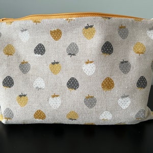Pode incluir: Uma bolsa retangular de tecido bege com um padrão repetido de morangos dourados, cinzentos e brancos. A bolsa tem um fecho de correr dourado. O tecido parece ser linho ou um material tecido semelhante. A bolsa é provavelmente usada para cosméticos ou pequenos objetos.