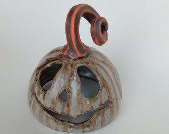 May include: A ceramic jack-o'-lantern with a smiling face and striped pattern. The pumpkin has a curved stem with orange and brown stripes. The pumpkin is a light gray color.