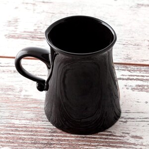 Puede incluir: Una taza de cer&aacute;mica negra con una base ancha y un asa redondeada. La taza est&aacute; vac&iacute;a y se encuentra sobre una superficie de madera blanca.