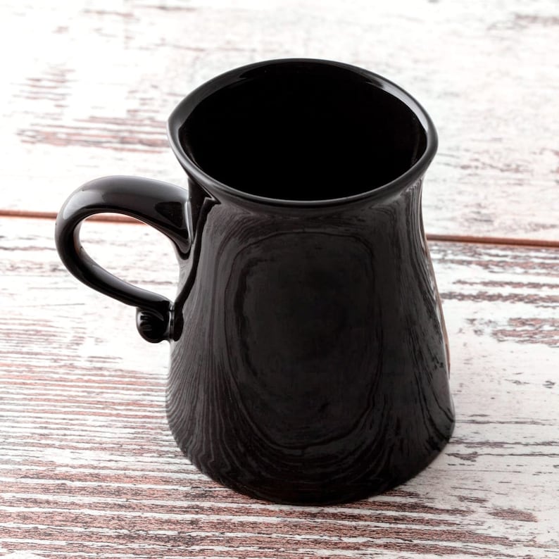 Puede incluir: Una taza de cer&aacute;mica negra con una base ancha y un asa redondeada. La taza est&aacute; vac&iacute;a y se encuentra sobre una superficie de madera blanca.
