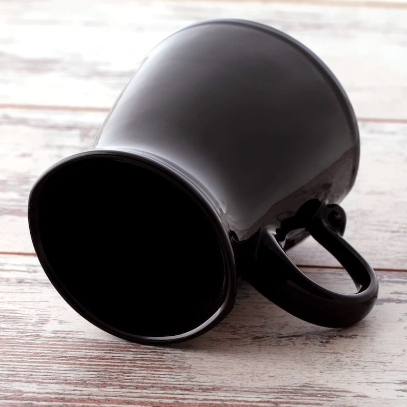 Puede incluir: Una taza de cer&aacute;mica negra con un asa ancha y redondeada. La taza est&aacute; sobre una superficie de madera blanca.