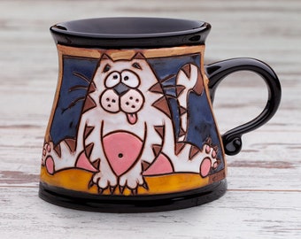 Puede incluir: Una taza de cerámica con borde y asa negros, que presenta una ilustración pintada a mano de un gato blanco con rayas marrones, vientre rosa y lengua rosa afuera, sobre un fondo azul oscuro y una base de color amarillo.