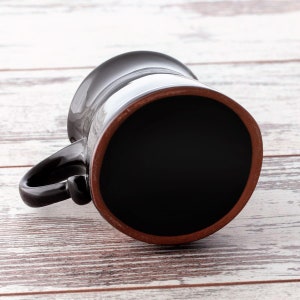Puede incluir: Una taza de cer&aacute;mica negra con un borde marr&oacute;n, vista desde abajo. La taza est&aacute; sobre una superficie de madera blanca.
