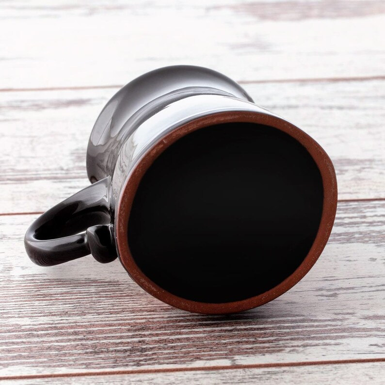 Puede incluir: Una taza de cer&aacute;mica negra con un borde marr&oacute;n, vista desde abajo. La taza est&aacute; sobre una superficie de madera blanca.