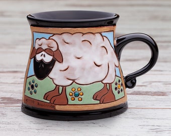 Puede incluir: Una taza de cerámica con borde y asa de color negro brillante, que presenta una ilustración pintada a mano de una oveja blanca con pezuñas y cara marrón sobre un fondo azul claro y verde. El texto gappa handmade está grabado abajo a la izquierda.