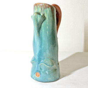 Puede incluir: Un jarrón de cerámica azul turquesa con una forma orgánica única y un asa marrón. El jarrón tiene una superficie texturizada y un pequeño sello naranja en la parte inferior.