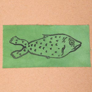 May include: A green fabric patch with a black fish illustration. The fish is depicted with a rounded body, spots, and a detailed eye. The rectangular patch is set against a tan background.