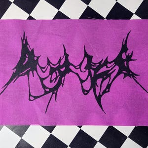 May include: A rectangular patch with a vibrant magenta background. A black, abstract design resembling a spiky, organic shape is centered on the patch. The patch is placed on a black and white checkered surface.
