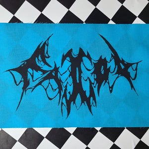 Op de afbeelding: Een blauwe stoffen patch met een zwart, gestileerd, gotisch logo. Het logo is een groot, abstract ontwerp met scherpe, gekartelde randen.