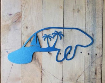 May include: A blue metal cutout of a manatee with palm trees inside the silhouette. The cutout is mounted on a wood background.
