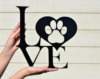 May include: Black metal sign spelling out the word "LOVE". The "O" is replaced with a heart shape containing a paw print. The sign is held up against a light-colored wooden background.