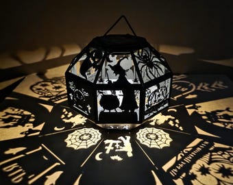 May include: A black Halloween lantern featuring cut-out designs of witches, spiders, and tombstones. The lantern casts spooky shadows of cobwebs, bats, and other Halloween-themed images onto the surface below. The lantern has a handle at the top.