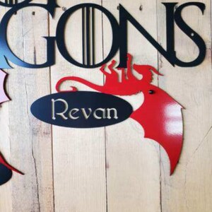 May include: A red metal dragon with a black oval sign that says "Revan" underneath. The dragon is attached to a wooden wall with the words "Game of Thrones" in black metal letters above it. The text "Dragon #2" is written below the dragon.