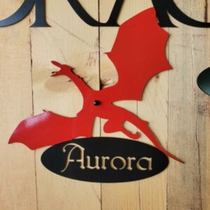May include: Red metal dragon silhouette with black metal wings and a black oval with the word "Aurora" in gold lettering. The dragon is mounted on a wooden surface. The text "Dragon #1" is below the dragon.